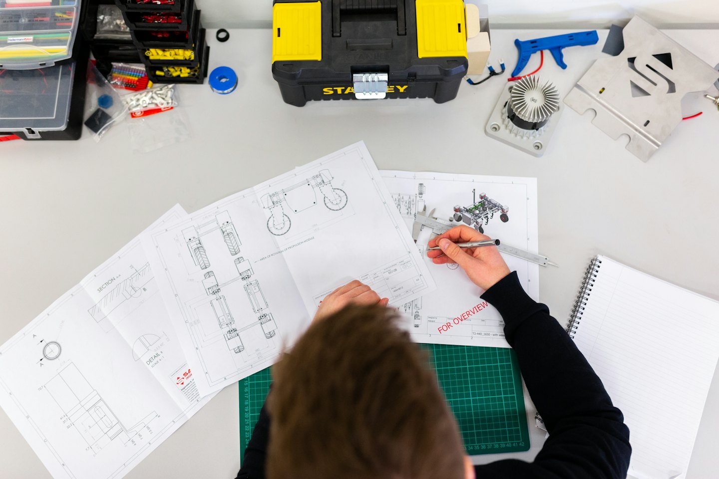 Engineer reviews schematics with calipers at a cluttered desk.
