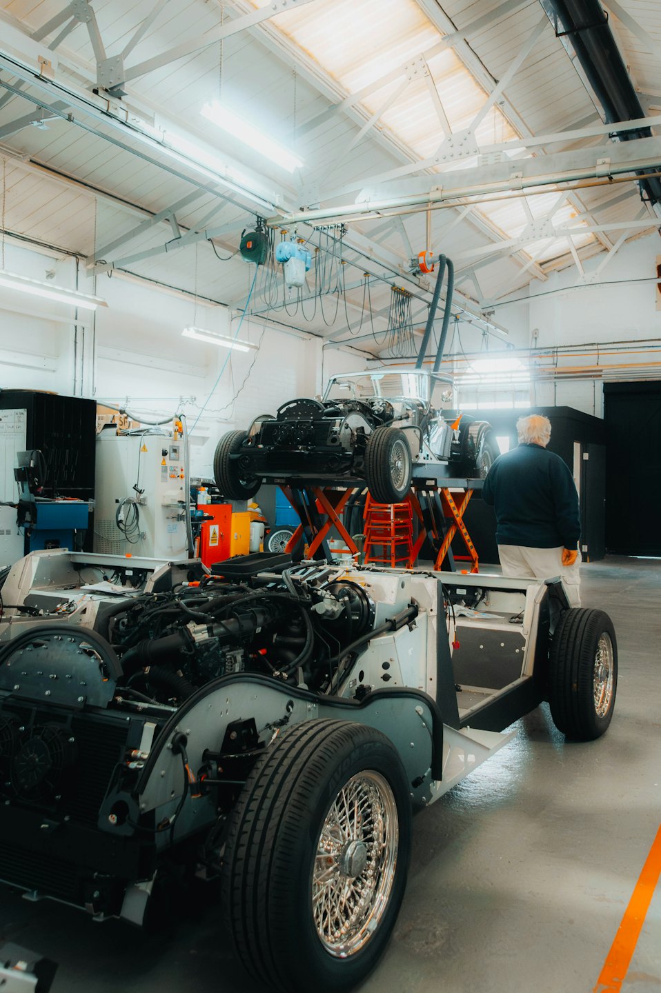 Man in workshop inspects white car chassis on lift.